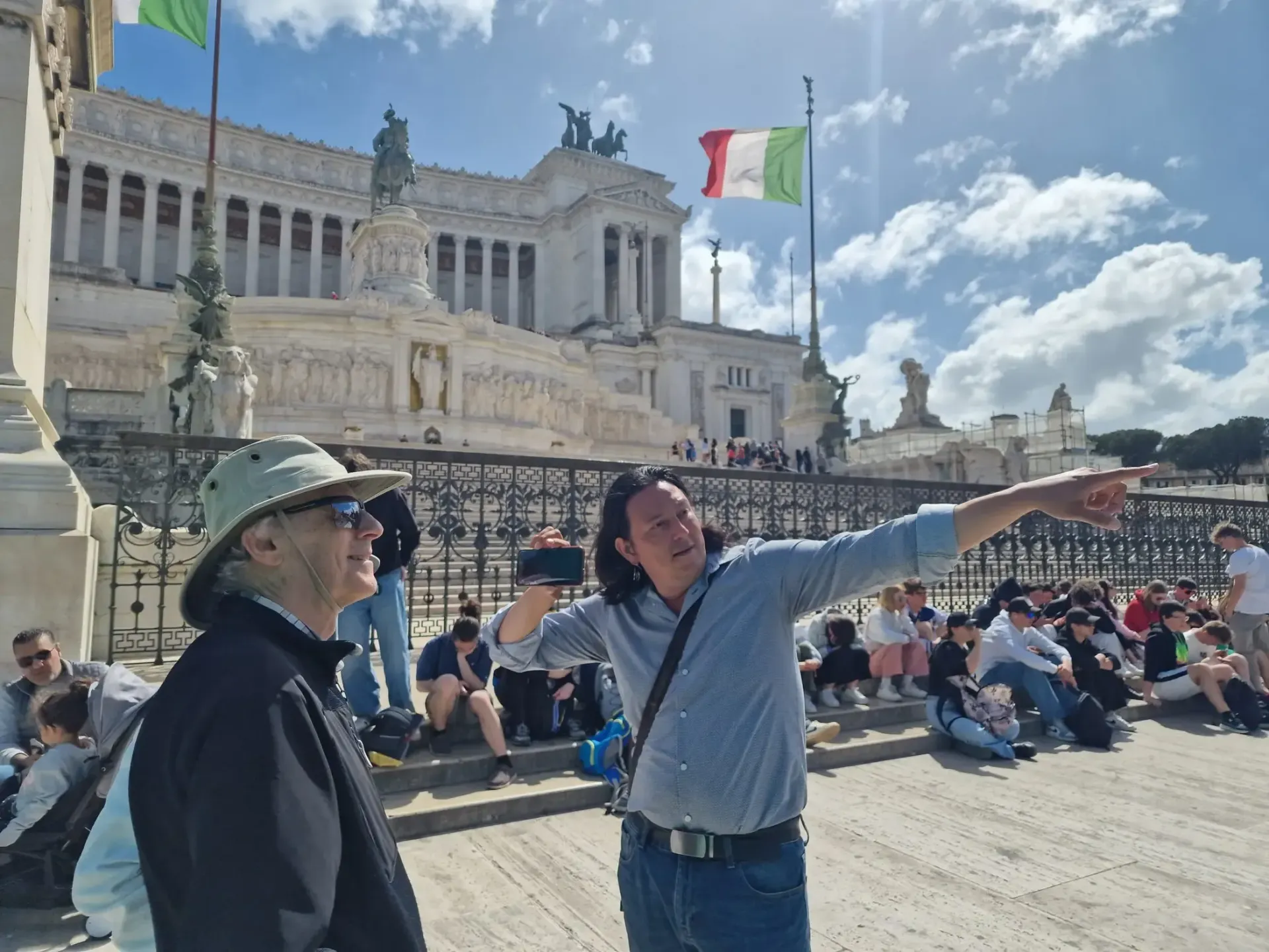 Man points towards a large white building, possibly giving a tour. Italian flag flies in the background. Sunny day.