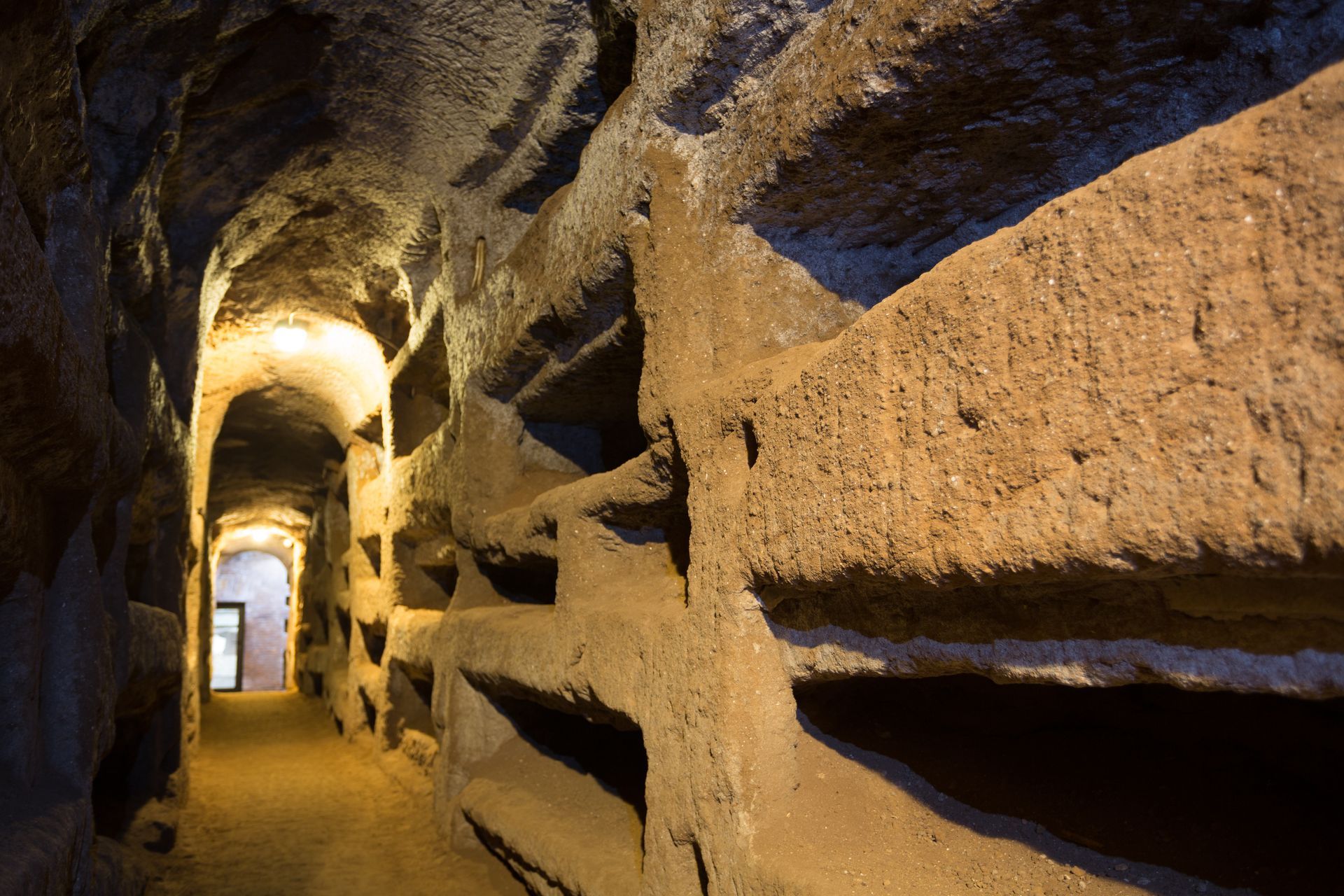 Tunnel lined with niches, possibly tombs, lit with warm light.
