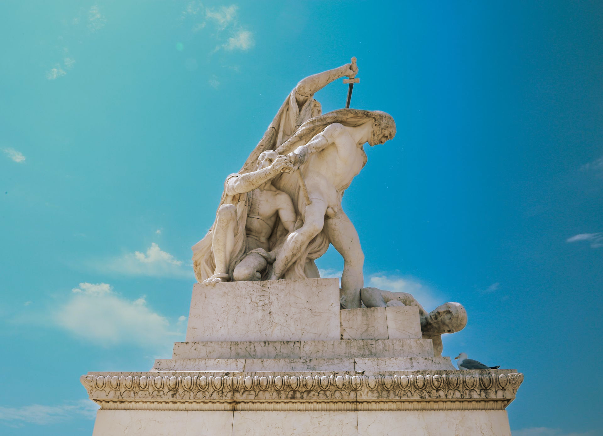 Sculpture of a man being stabbed by another figure; blue sky background.