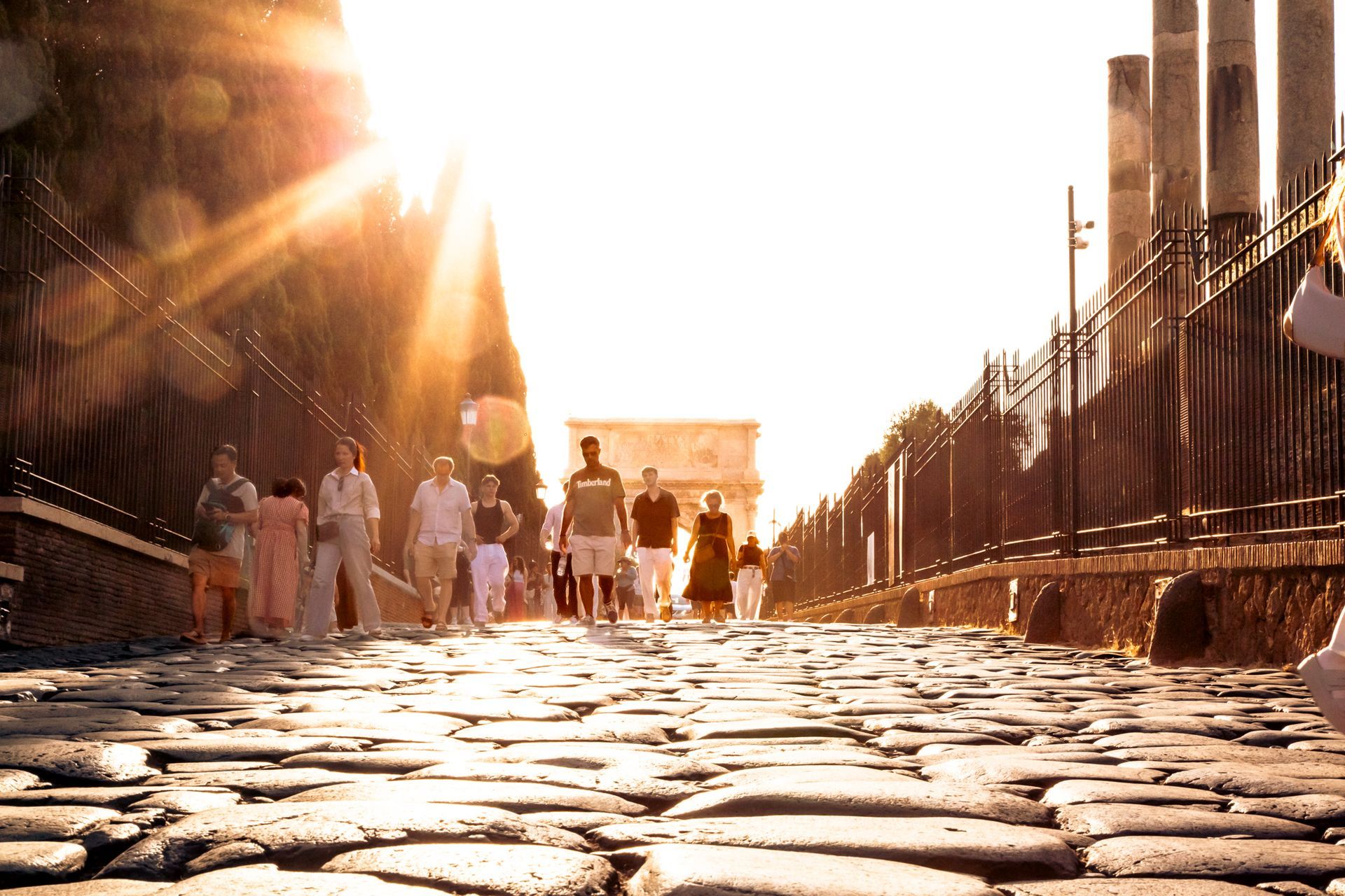 Cobblestone road in Rome with people walking towards the Arch of Constantine, sunburst in the background.