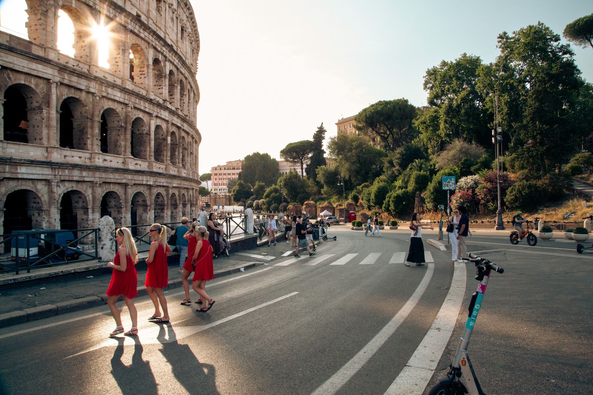 Colosseum in Rome, Italy, with people crossing street. Sun shines brightly. Some are wearing red dresses.