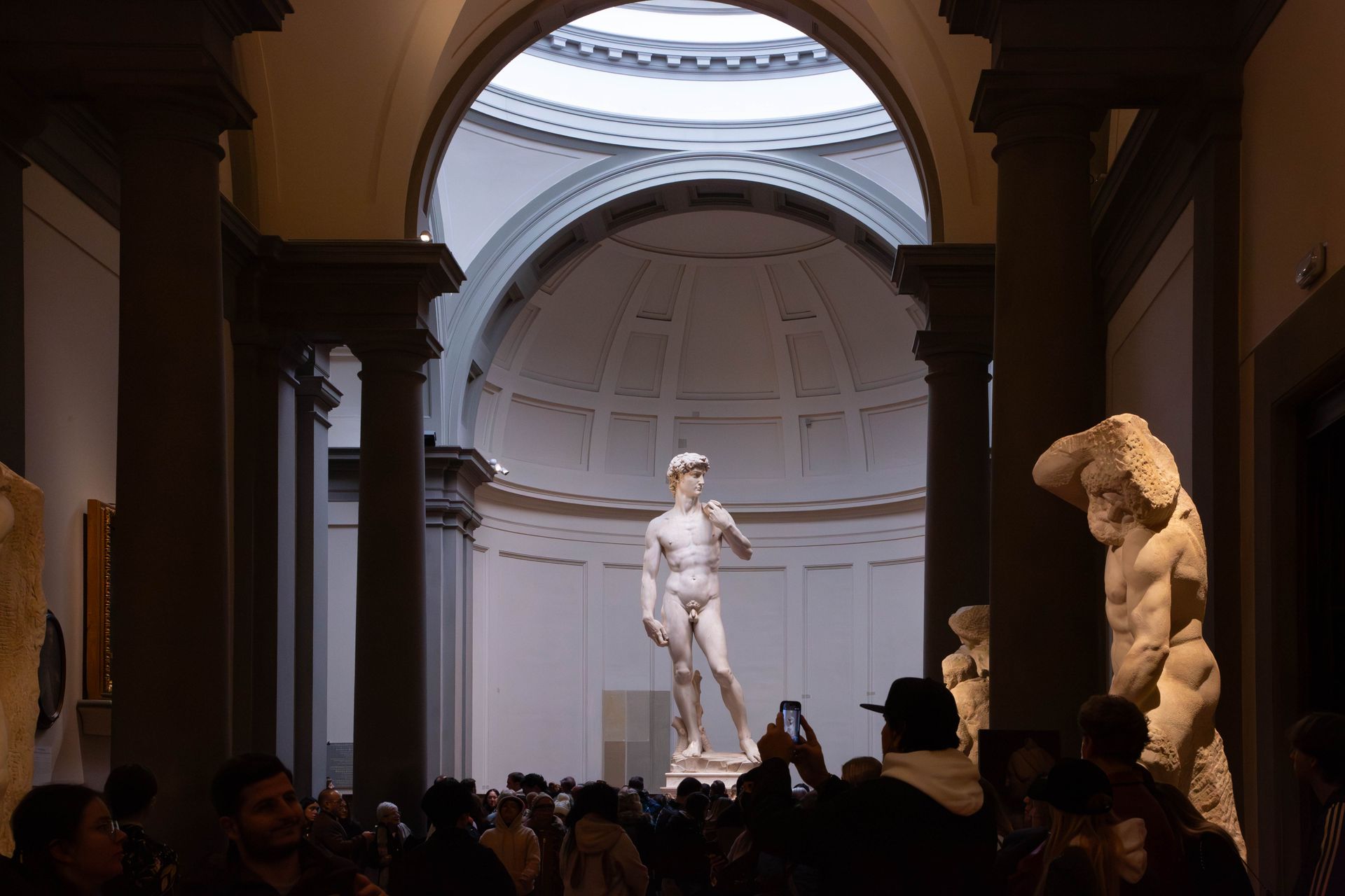 Classical statue in a museum hall, seen through an arched doorway, with visitors in silhouette below.