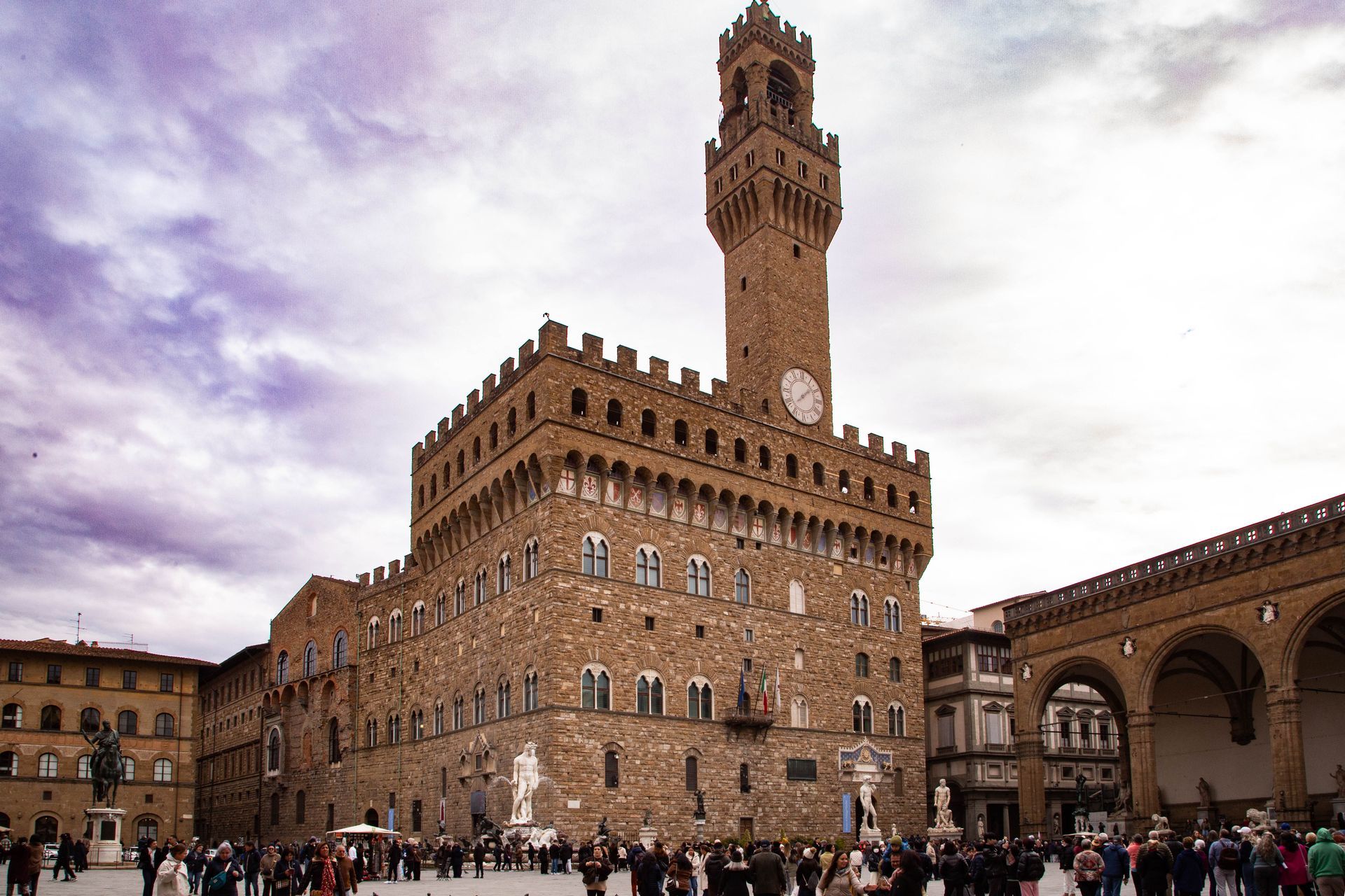 Historic brick palace with a tall clock tower and crowds in a public square under a cloudy sky