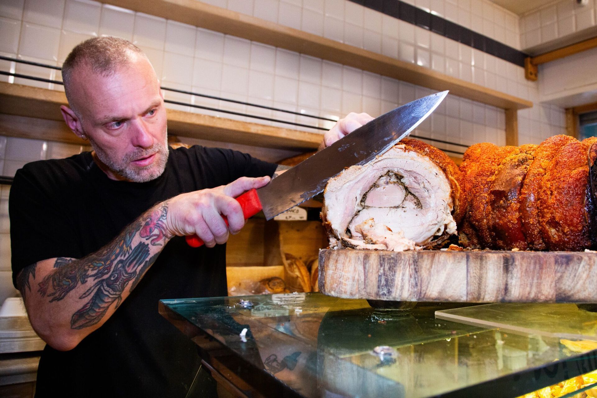 Man in blue shirt, cutting cheese at a market stall. Smiling, with people and produce in background.