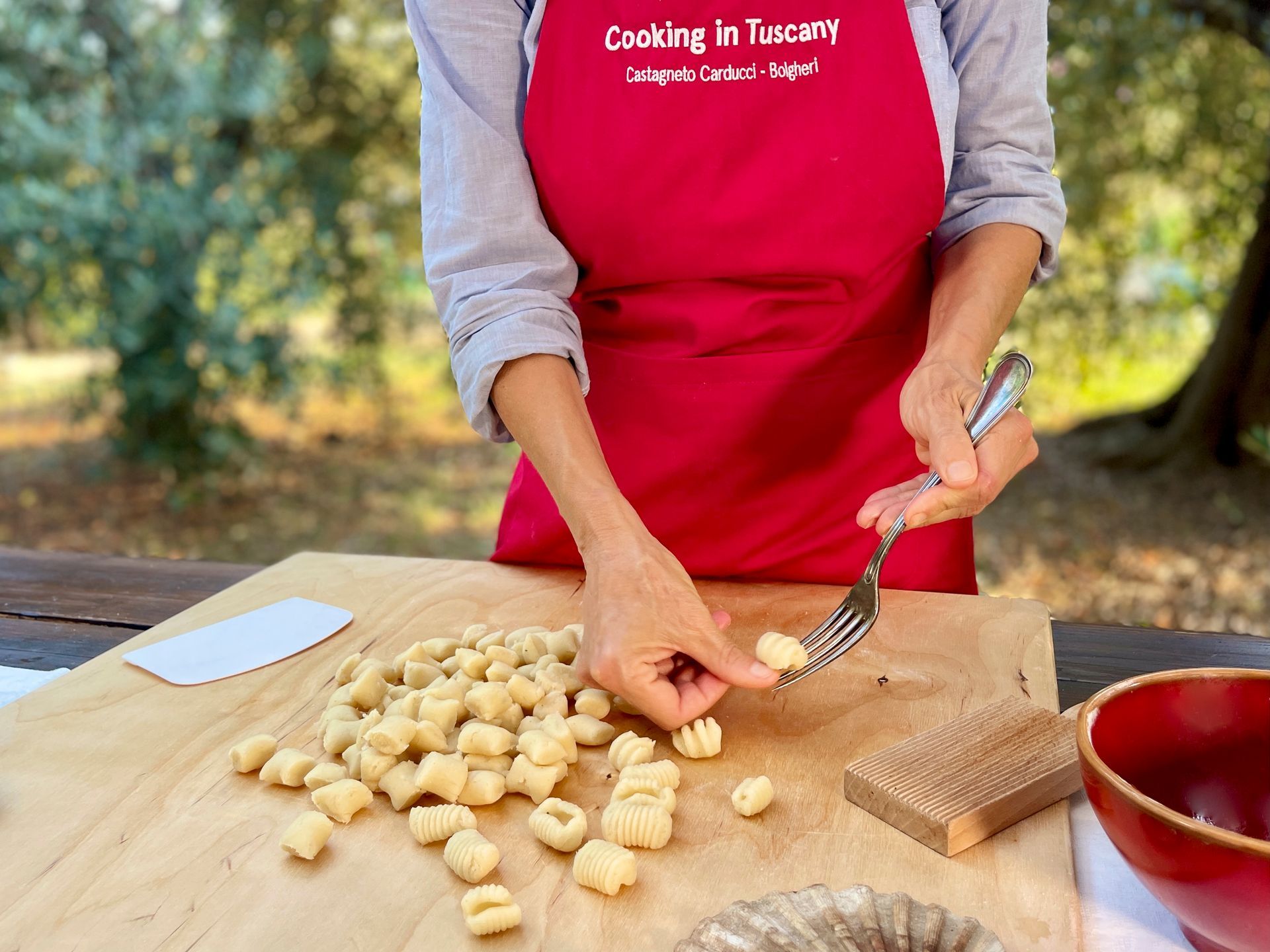 Person in a red apron shaping dough gnocchi on a wooden board outdoors.