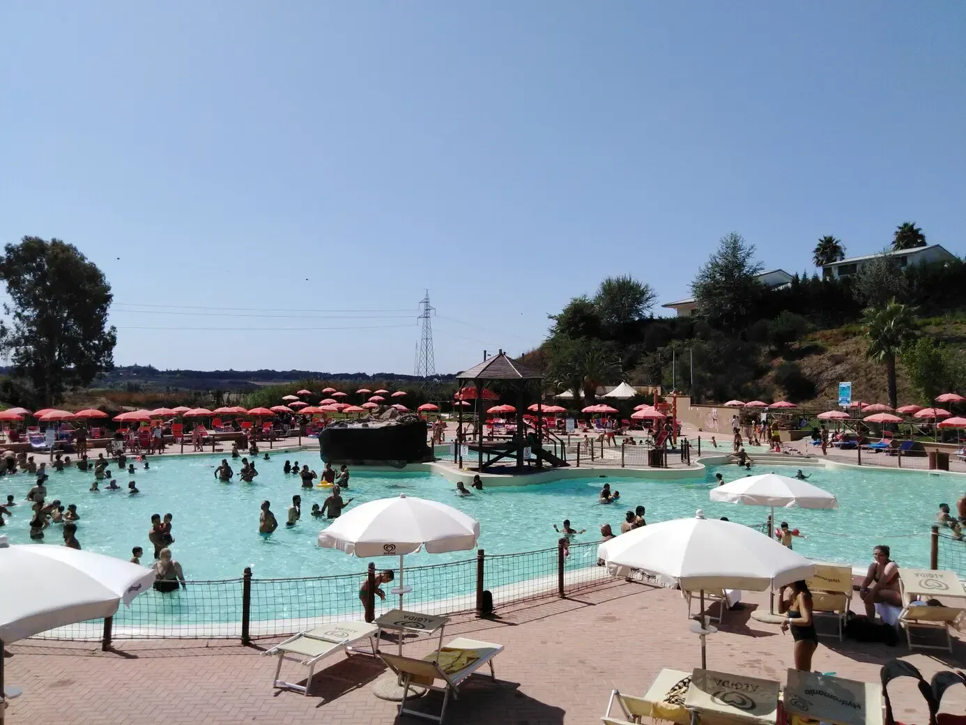 Large pool filled with people, surrounded by umbrellas and lounge chairs, under a clear sky.