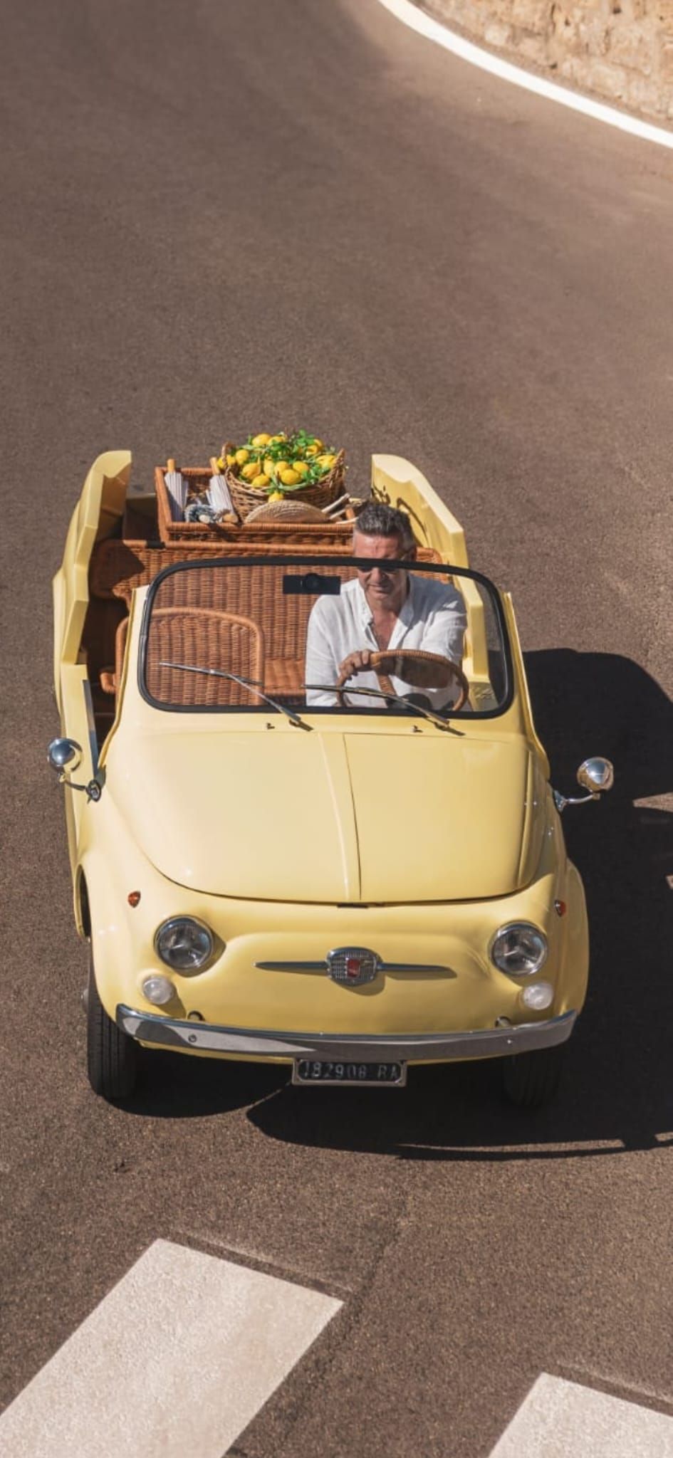Yellow vintage Fiat 500 driving on a curving road, with a plant-filled roof rack.