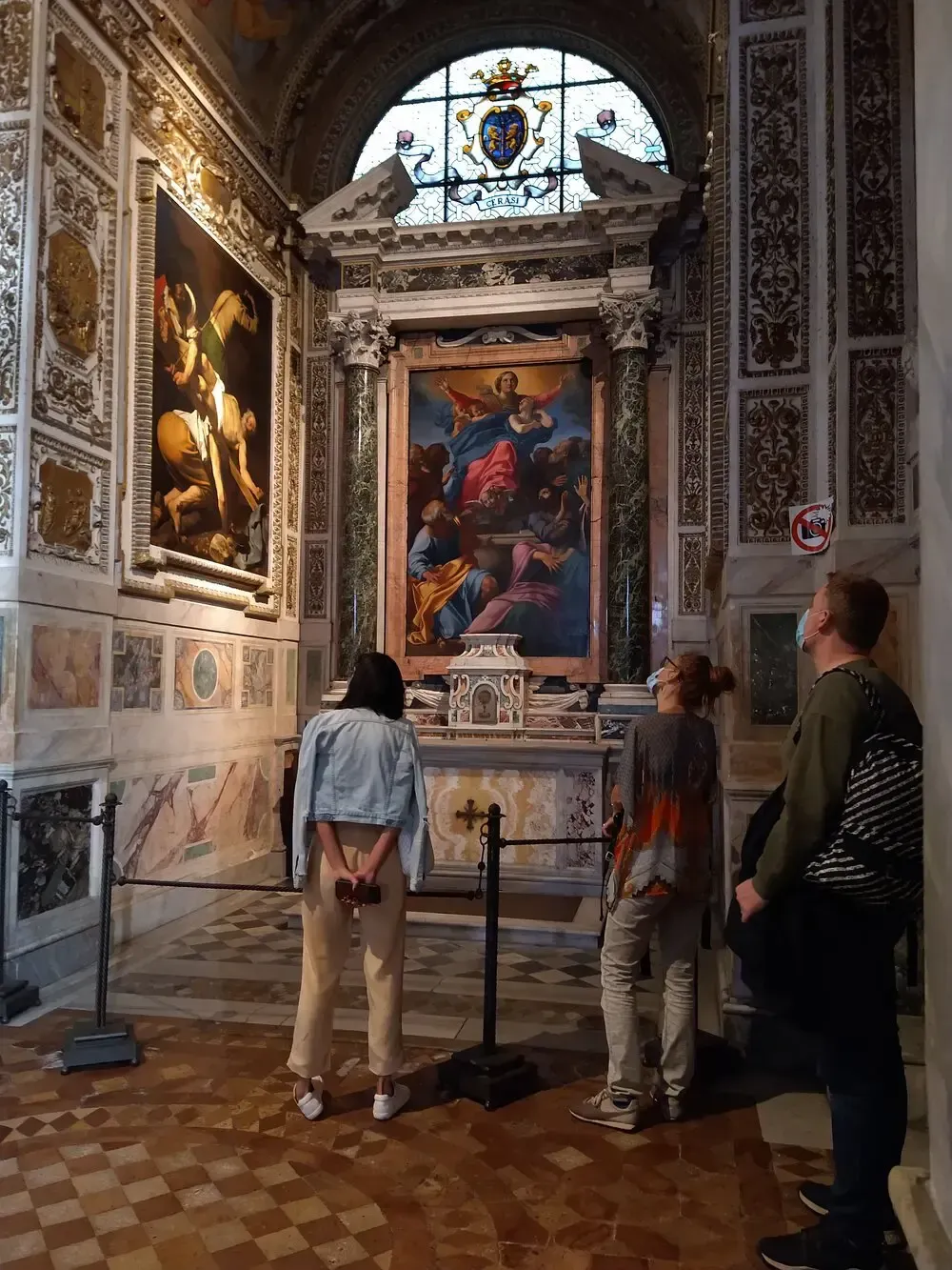 People in a church viewing a painting in an ornate alcove. Marble walls, stained glass, and a painting of figures.