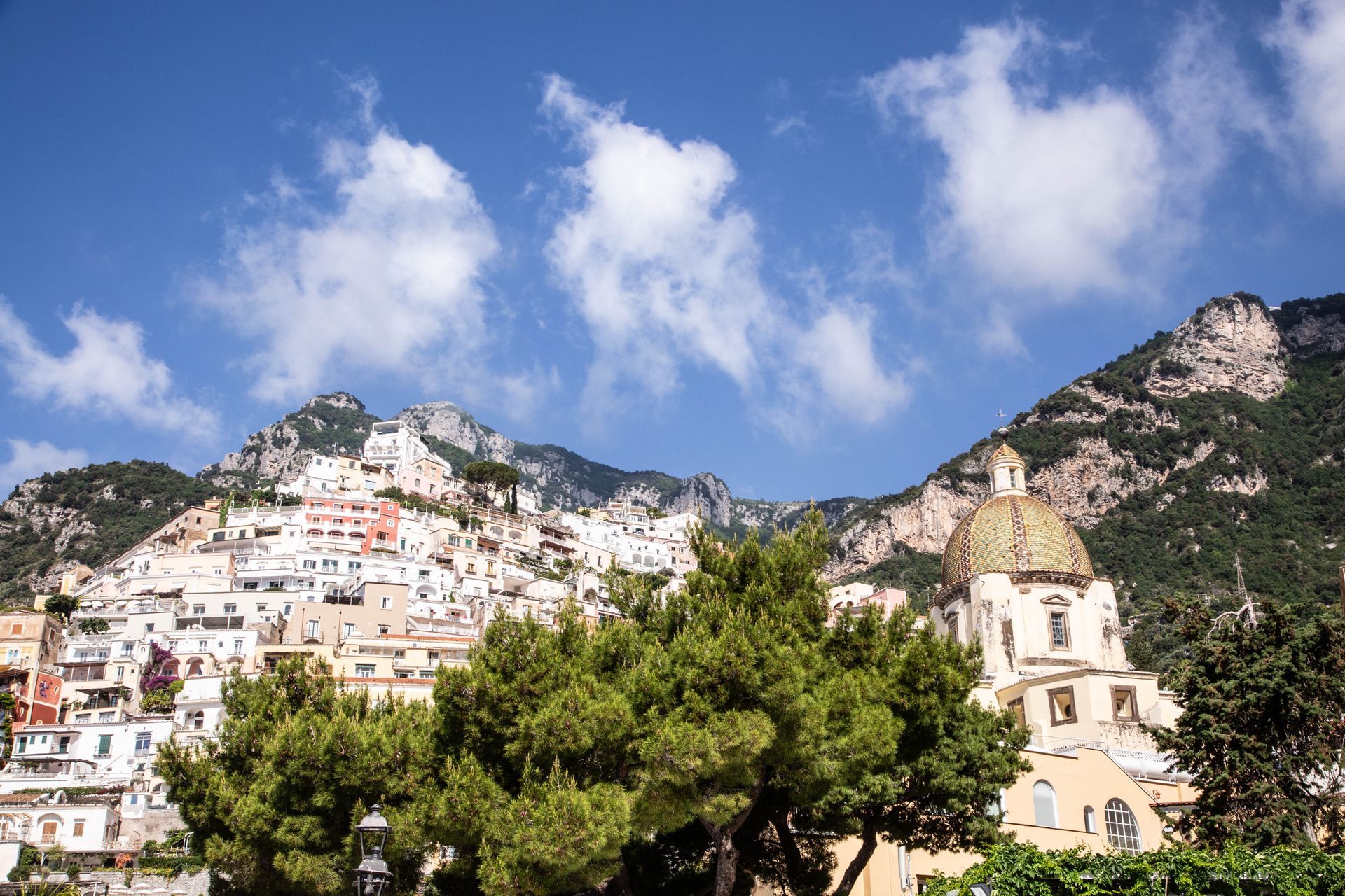 White hillside village beneath rocky mountains and blue sky, with trees and a domed church in the foreground