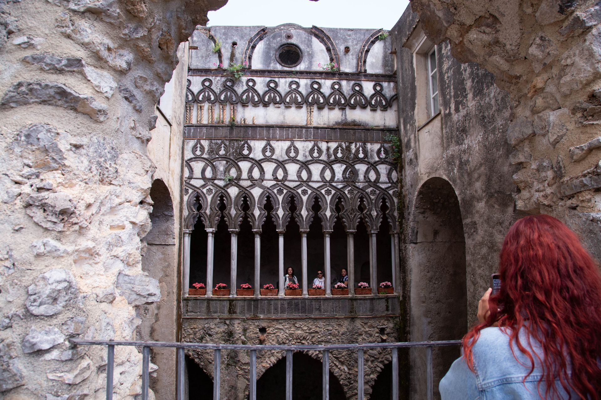 Woman with red hair looking through an ornate iron gate in a stone archway