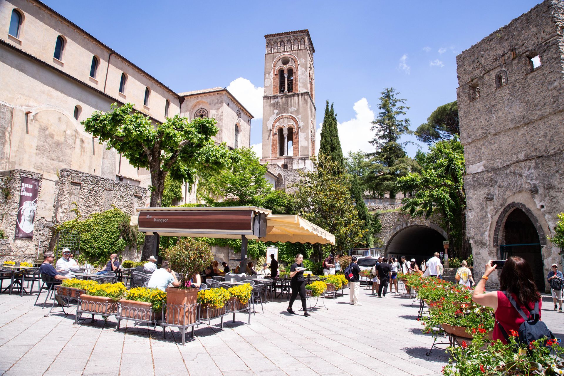 Sunny plaza café in a stone courtyard with flowers and a tall historic tower in the background