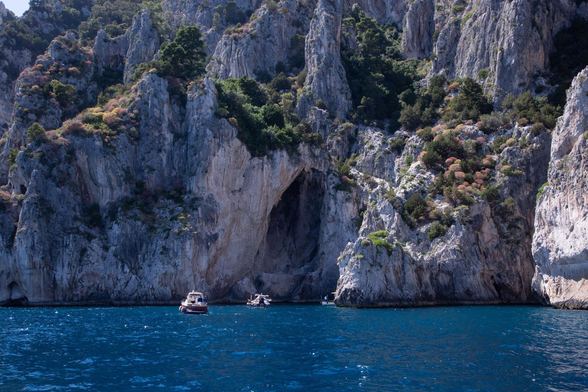 Rocky seaside cliffs with a cave above bright blue water and two small boats below