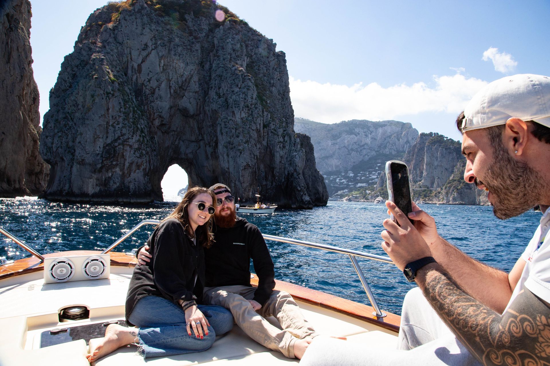 Two people on a boat taking a selfie near a rocky sea arch on a sunny day