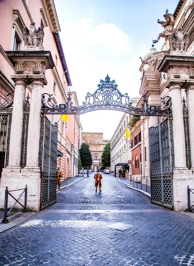 Ornate gate frames a cobblestone street in Rome. Person stands in center. Buildings line both sides.