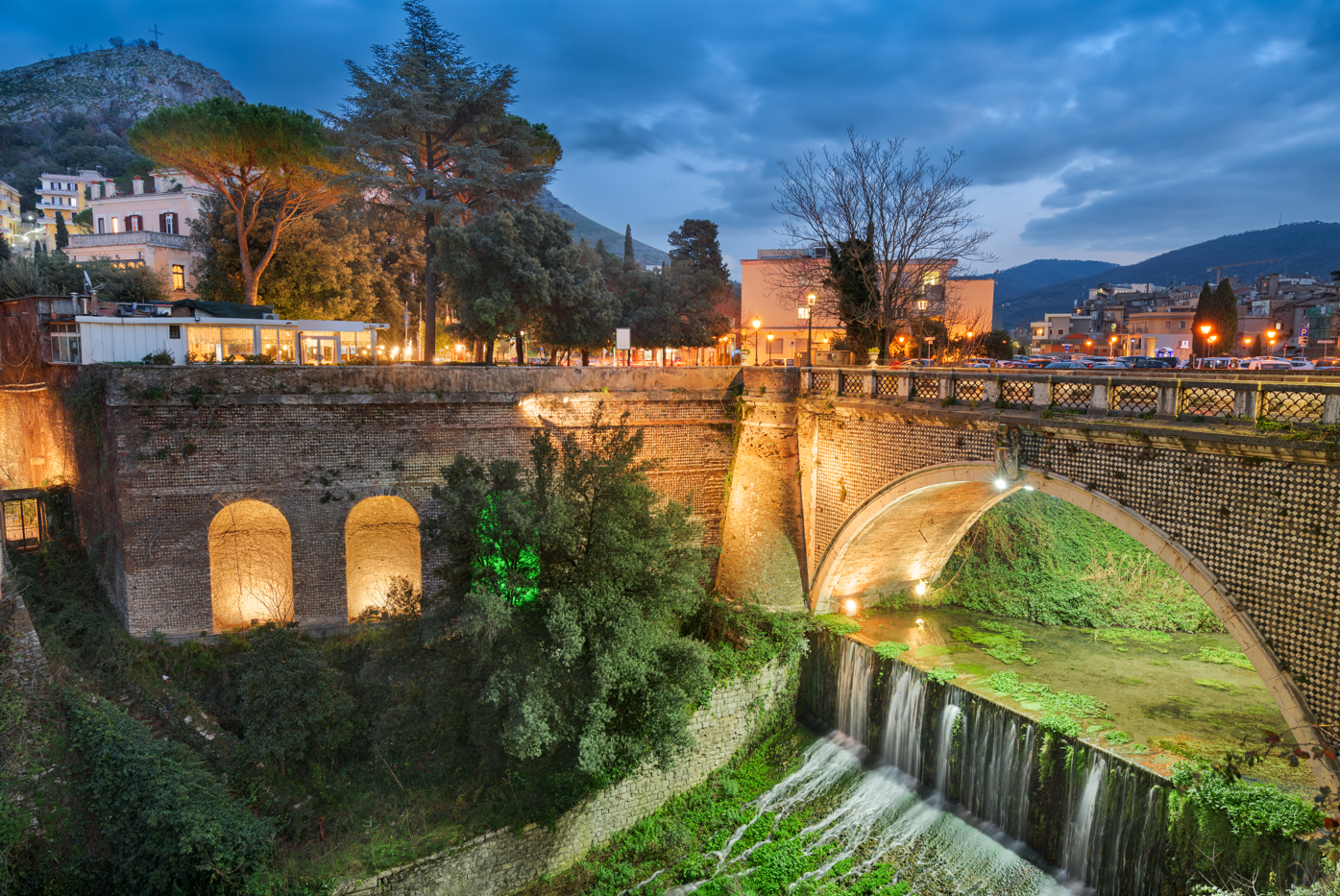 Stone bridge over a waterfall at dusk, with buildings and mountains in the background, illuminated by lights.