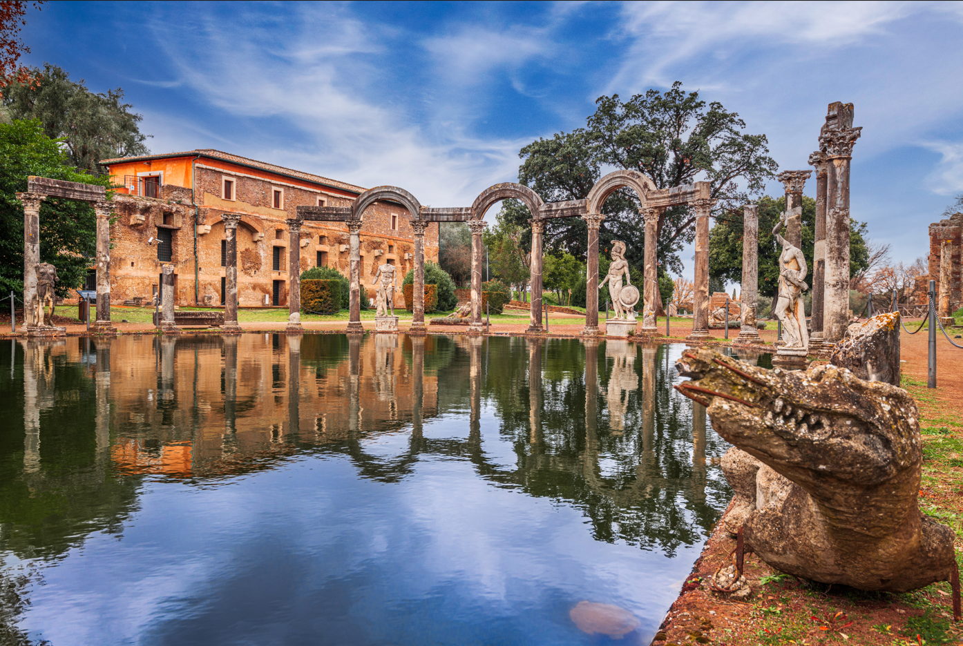 Ancient Roman ruins reflected in water. Buildings, columns, statues in a park-like setting.