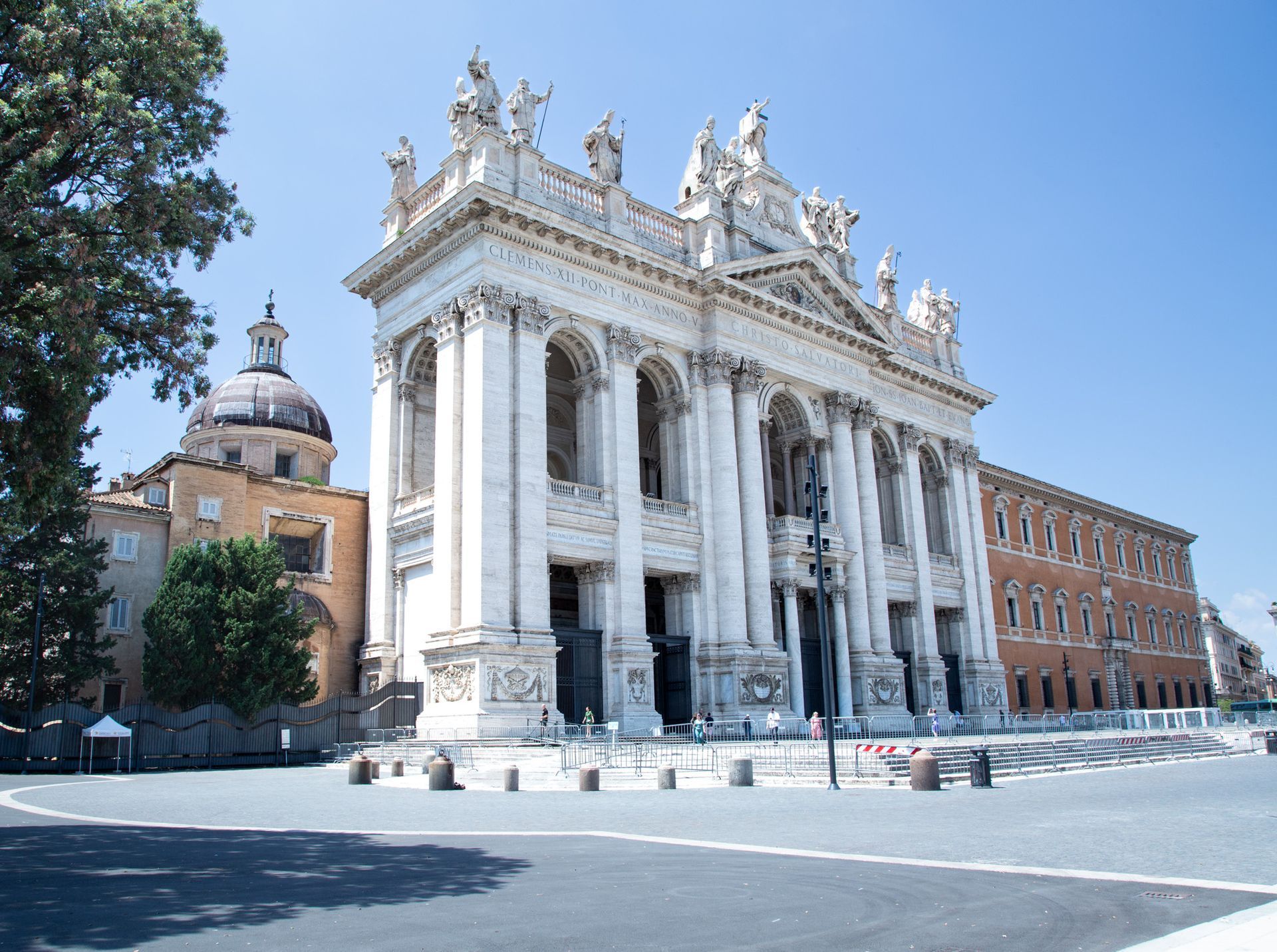 Grand white marble church facade with columns and statues beside a brick building on a sunny street