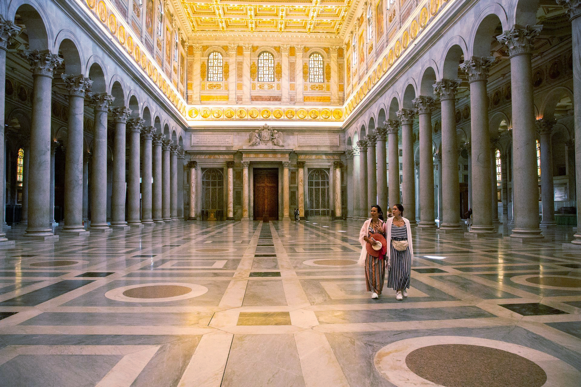 Grand marble hall with gilded ceiling and two people walking across the patterned floor