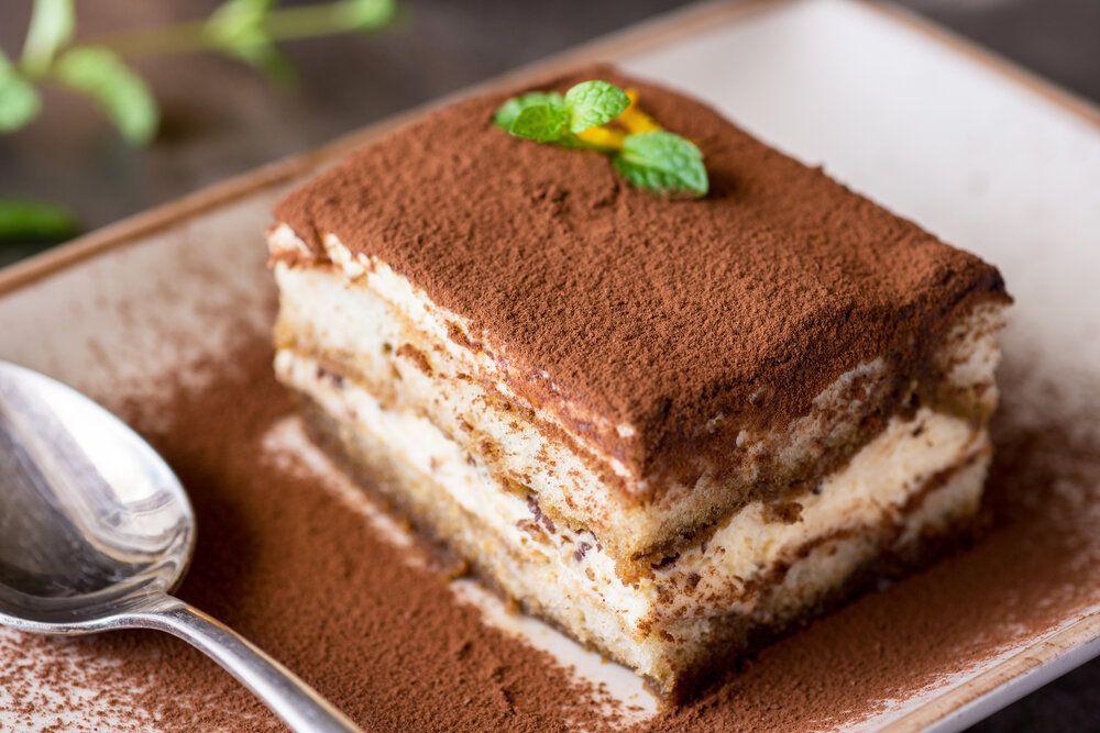 Tiramisu dessert on a square plate, dusted with cocoa powder and topped with a mint sprig, with a spoon alongside.