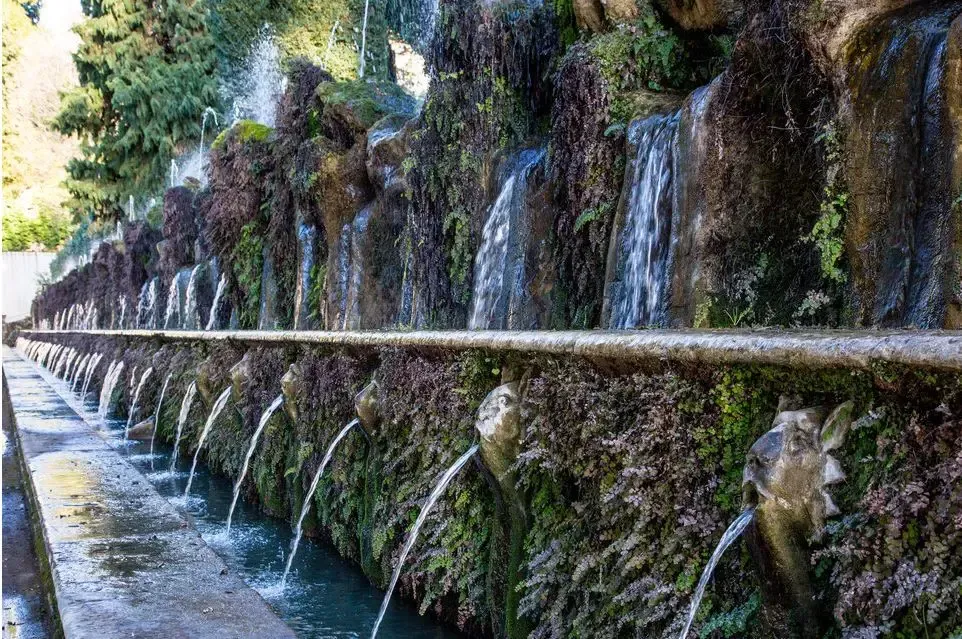 Stone fountain with multiple spouts cascading water into a trough; lush greenery.