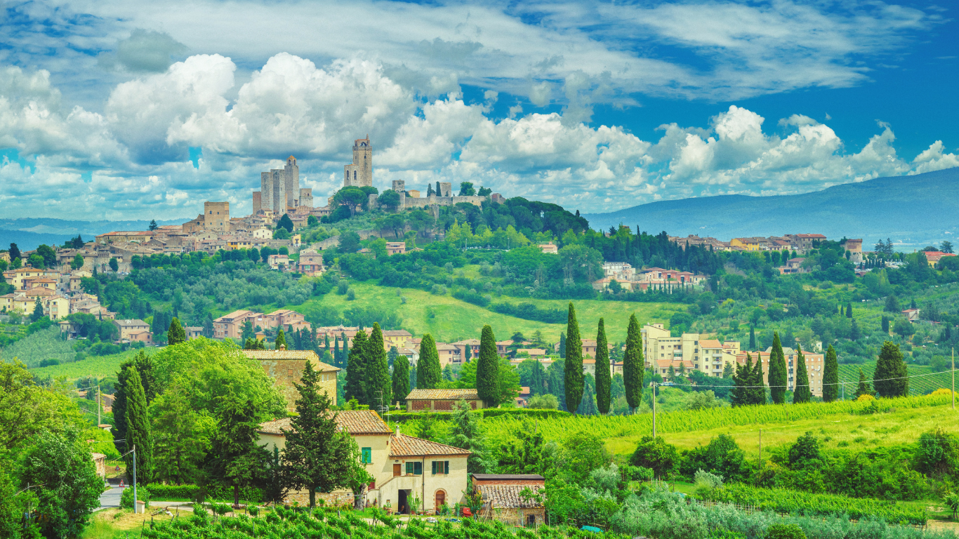 Hilltop city with historic towers above lush green landscape under a bright blue, cloudy sky