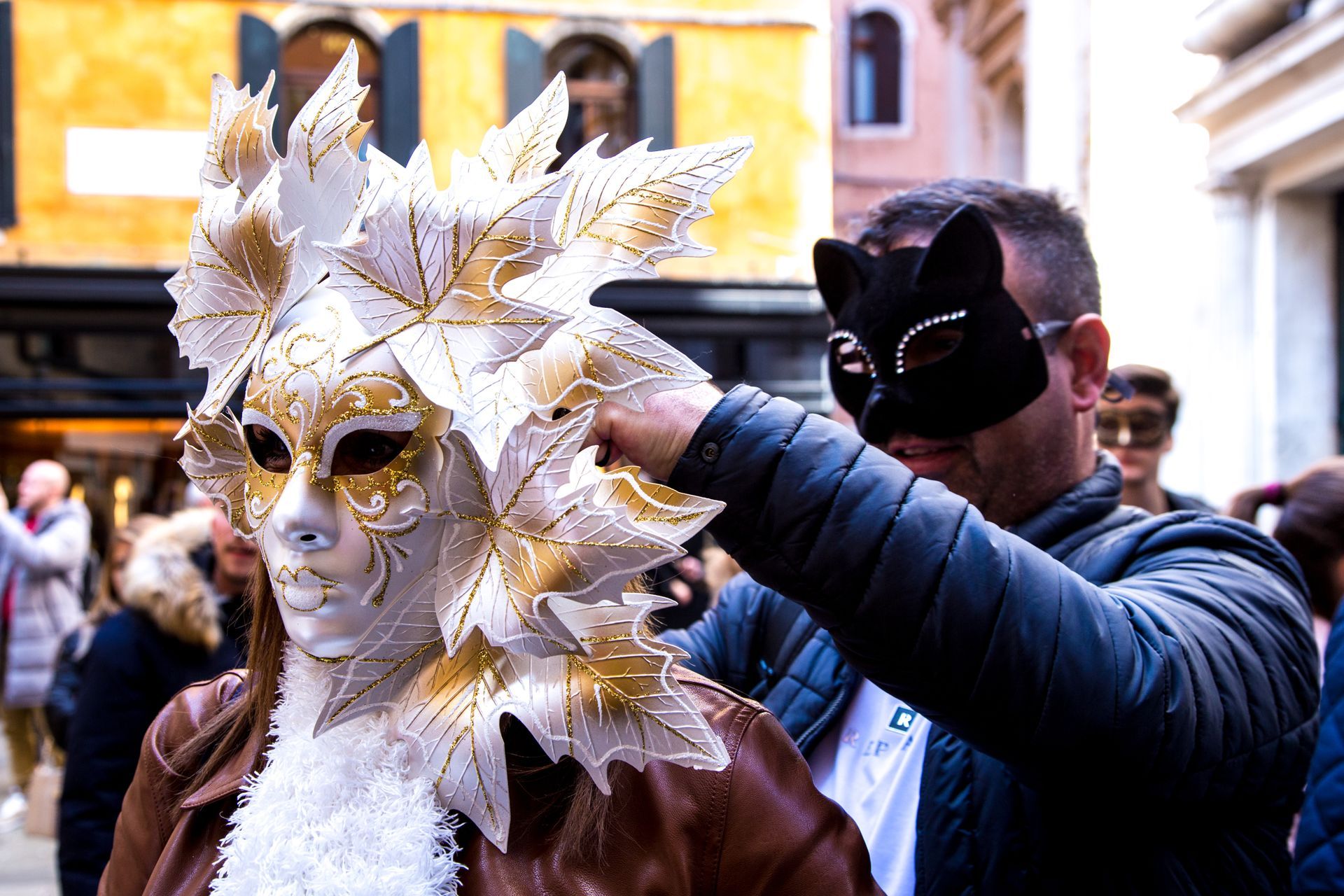 Masked performer in gold-and-white feathered headdress being adjusted by another person on a street