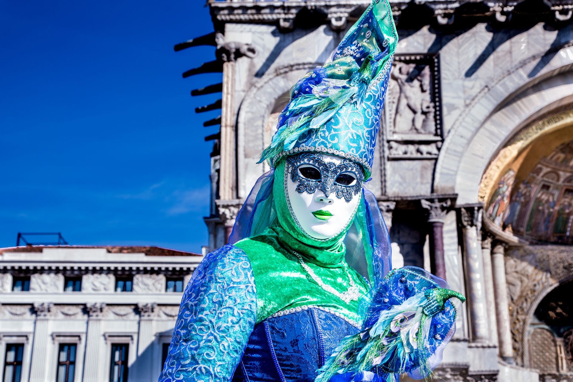 Blue-and-green costumed street performer with a tall hat and mask in a sunlit plaza