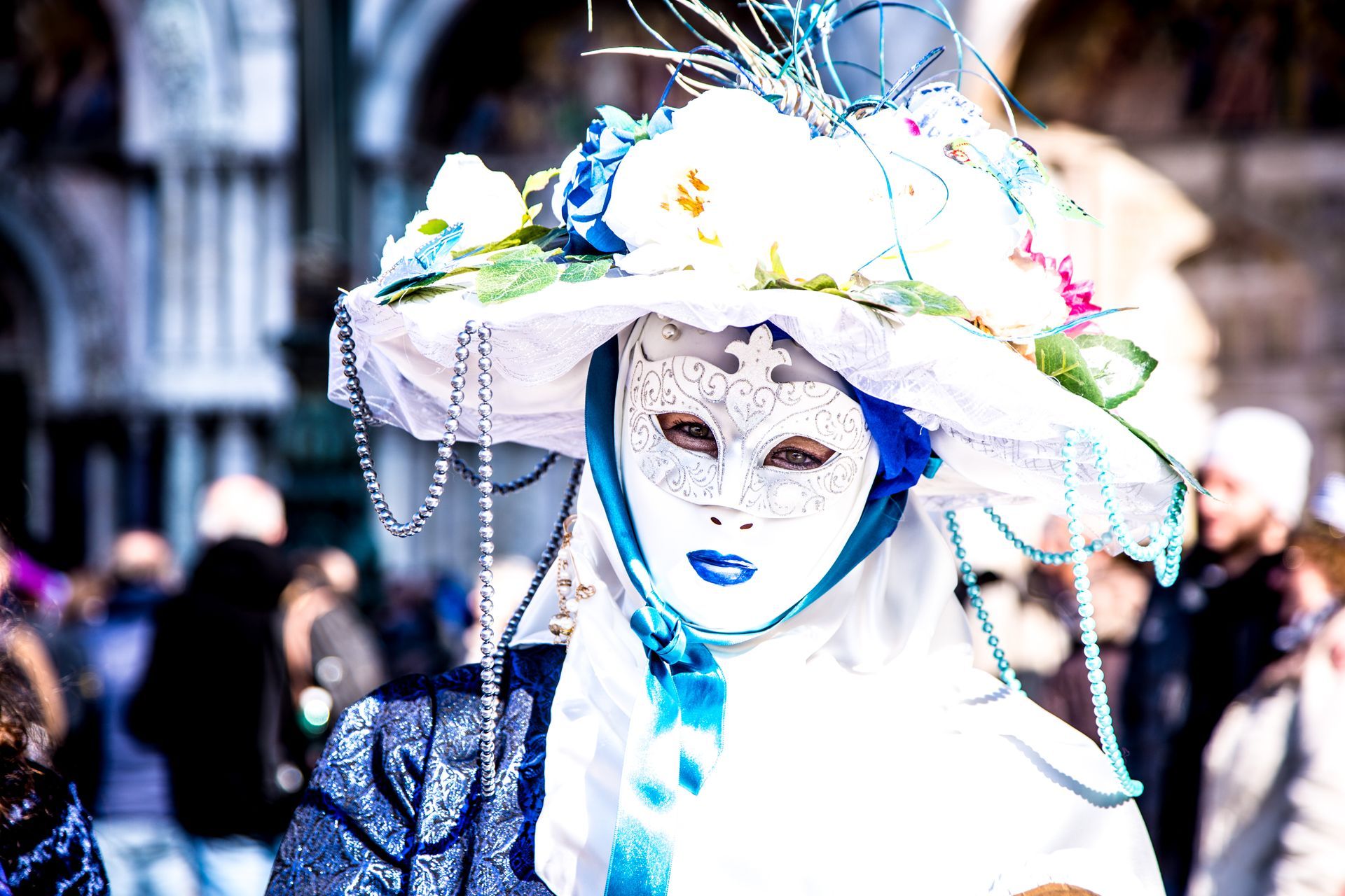 Person in a white lace mask and floral hat at a colorful street festival