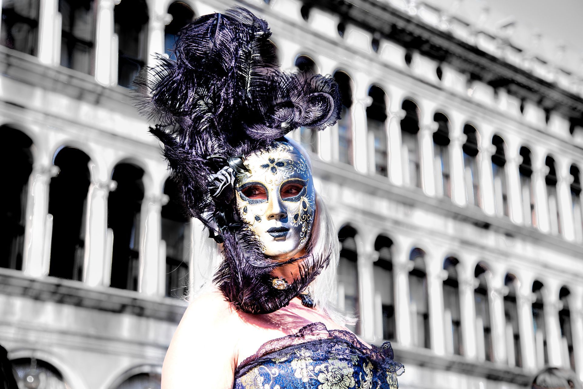 Person in ornate blue and black costume with dramatic headpiece, standing before a white historic building.
