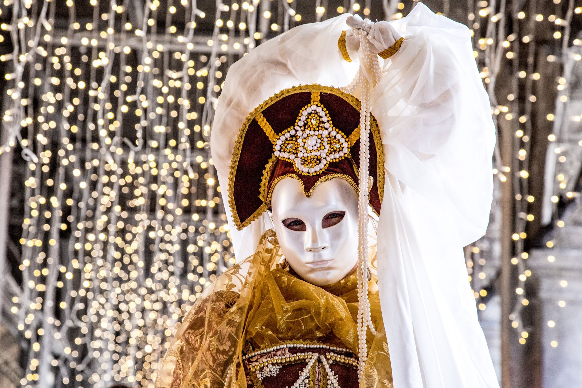 Masked figure in ornate gold costume before a curtain of warm string lights