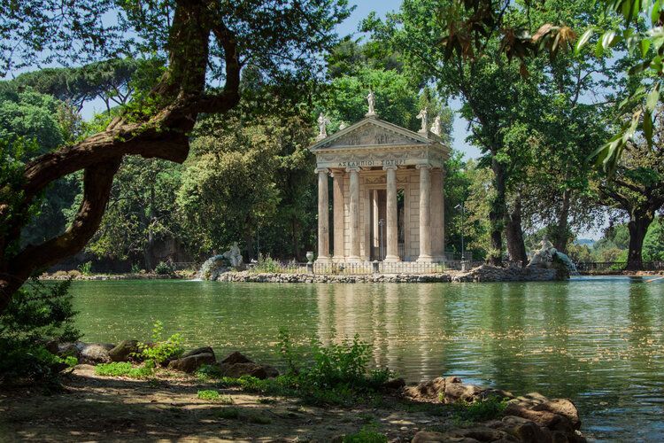 Temple of Asclepius on an island in a lake surrounded by trees; sunny day.