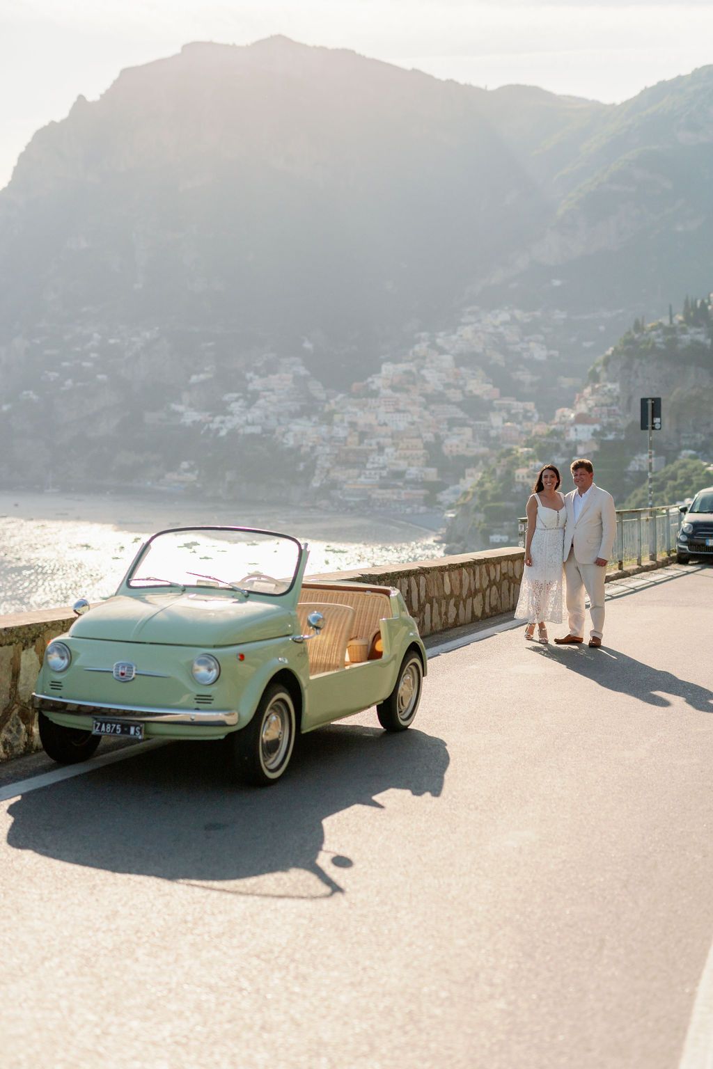 Vintage green Fiat 500 parked by a lakeside road, with two people walking beside a mountain backdrop.
