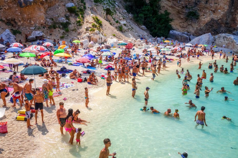 Beach crowded with people, colorful umbrellas, turquoise water, rocky cliffs.