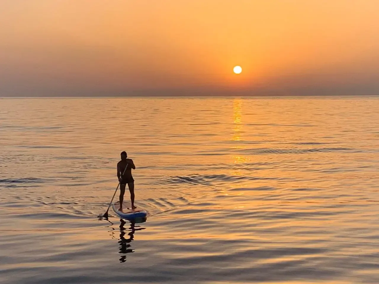 Person paddleboards on calm water toward the sunset.