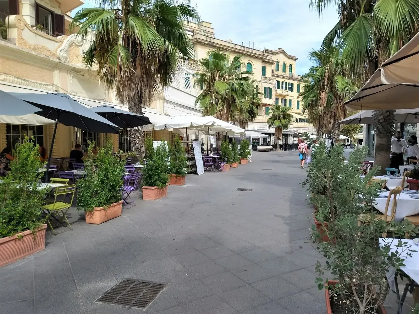 A pedestrian street lined with restaurants, outdoor seating, trees, and a building in the distance.