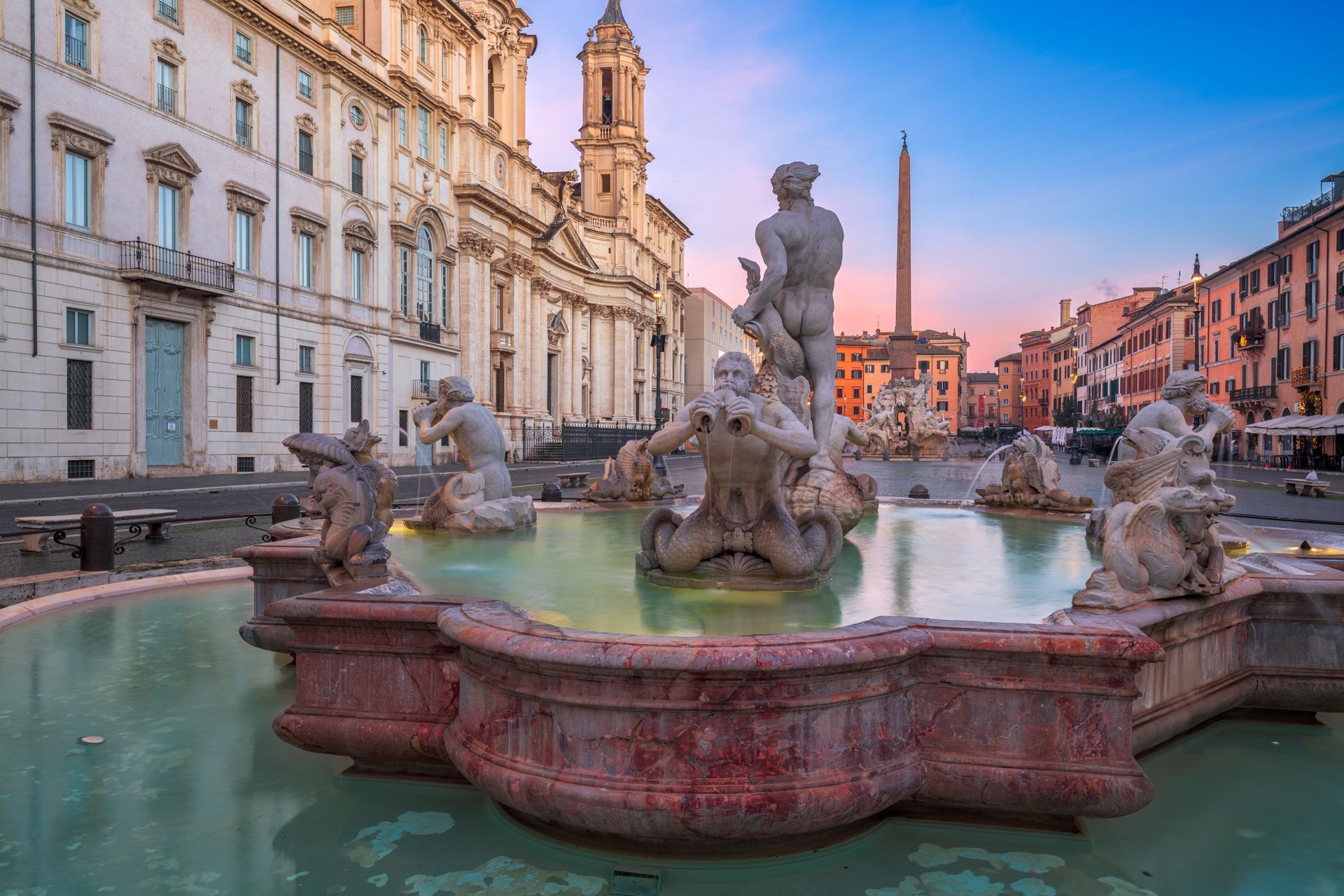 Fountain of Four Rivers in Piazza Navona, Rome, featuring sculptures and an obelisk; buildings in background at dawn.