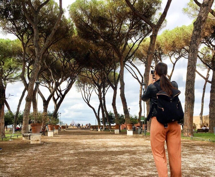 Person photographing a tree-lined path. They wear an orange pant, black jacket, and backpack. A cloudy sky is overhead.