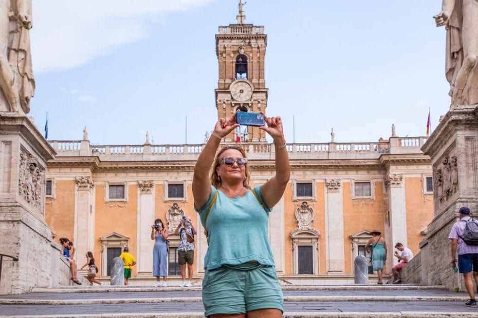 Woman taking a photo with a smartphone in front of a clock tower and a building with stairs in Rome.