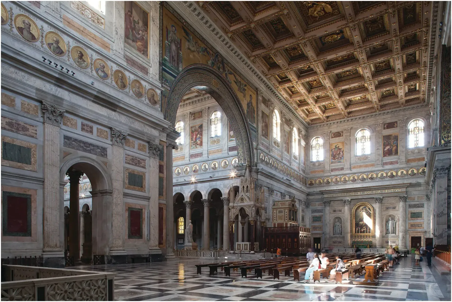 Interior of a large basilica with ornate marble walls, columns, and a coffered ceiling. Sunlight streams through windows.