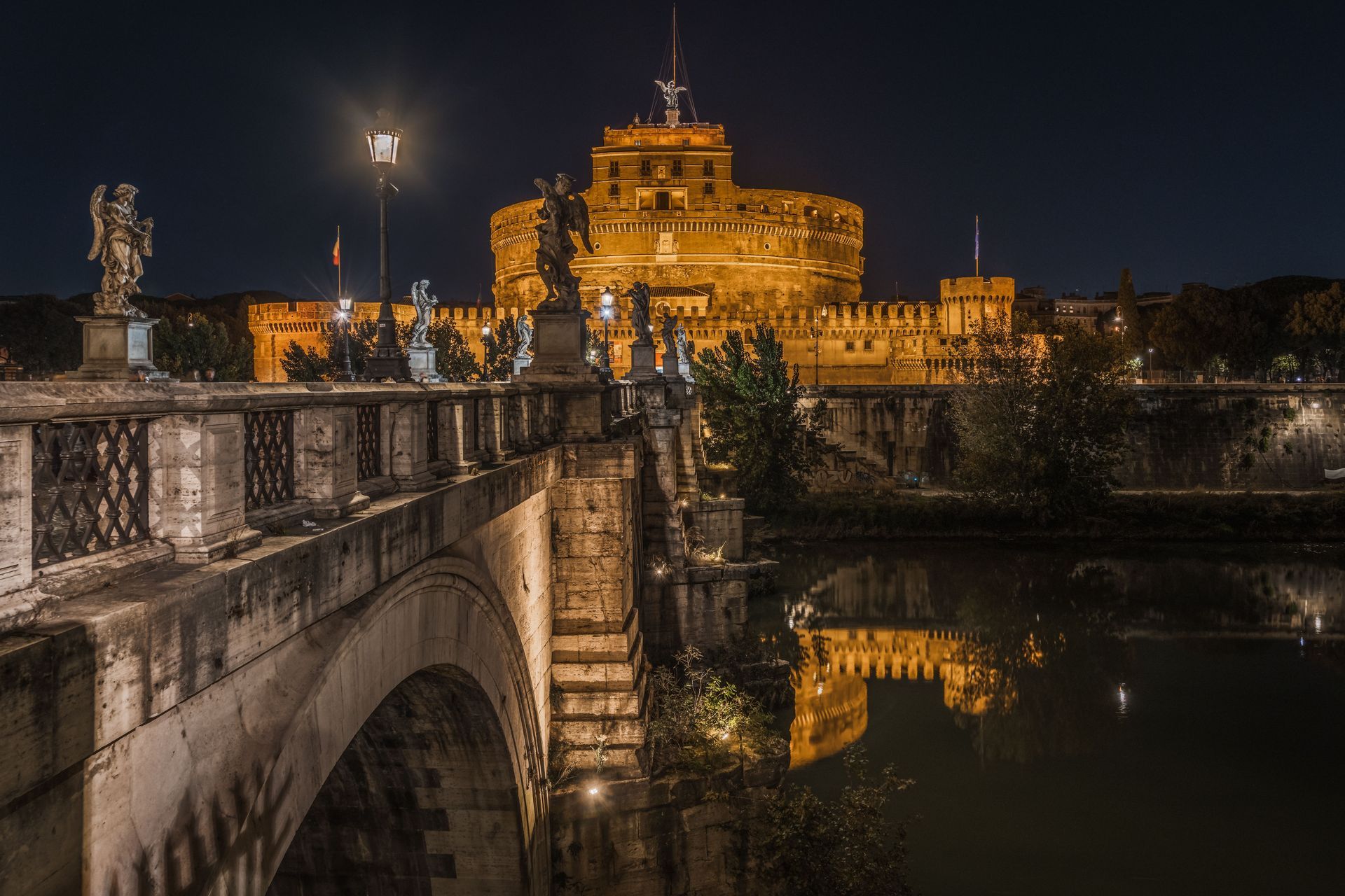 Night view of Castel Sant'Angelo in Rome, Italy, lit up, reflected in the Tiber River, with a bridge in foreground.
