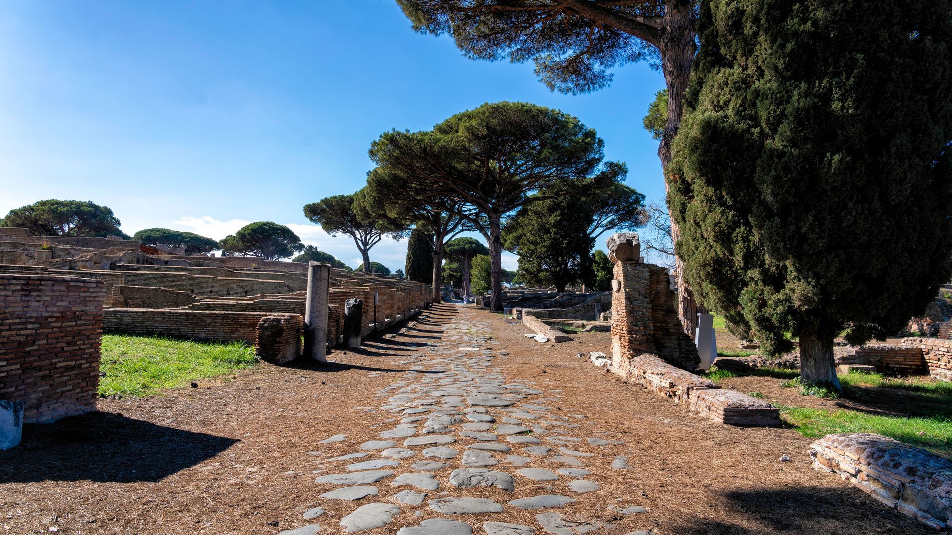 Ancient Roman cobblestone road lined with ruins and trees under a blue sky.