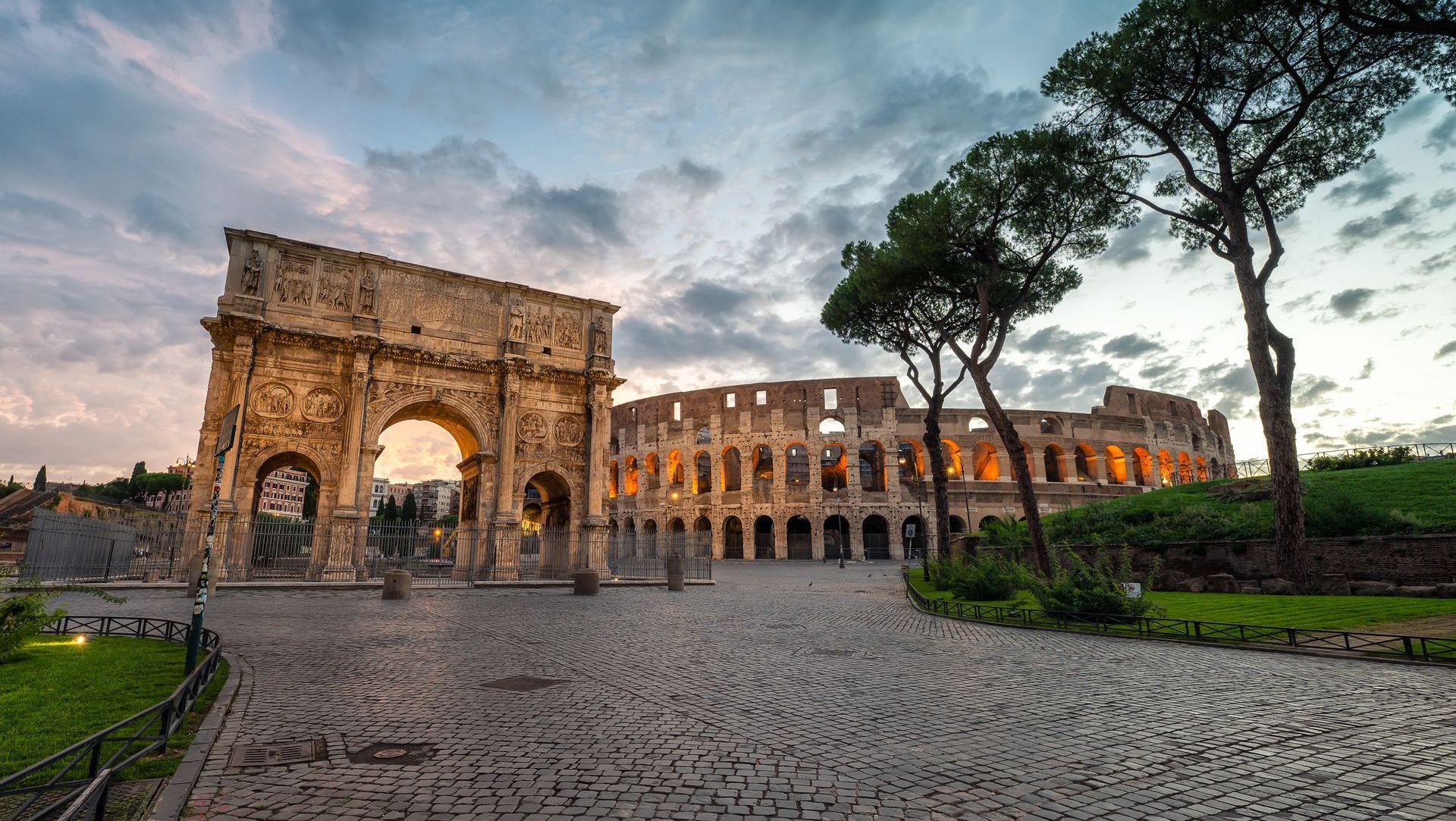 Arch of Constantine and Colosseum in Rome at dusk, cobbled road, trees, and cloudy sky.