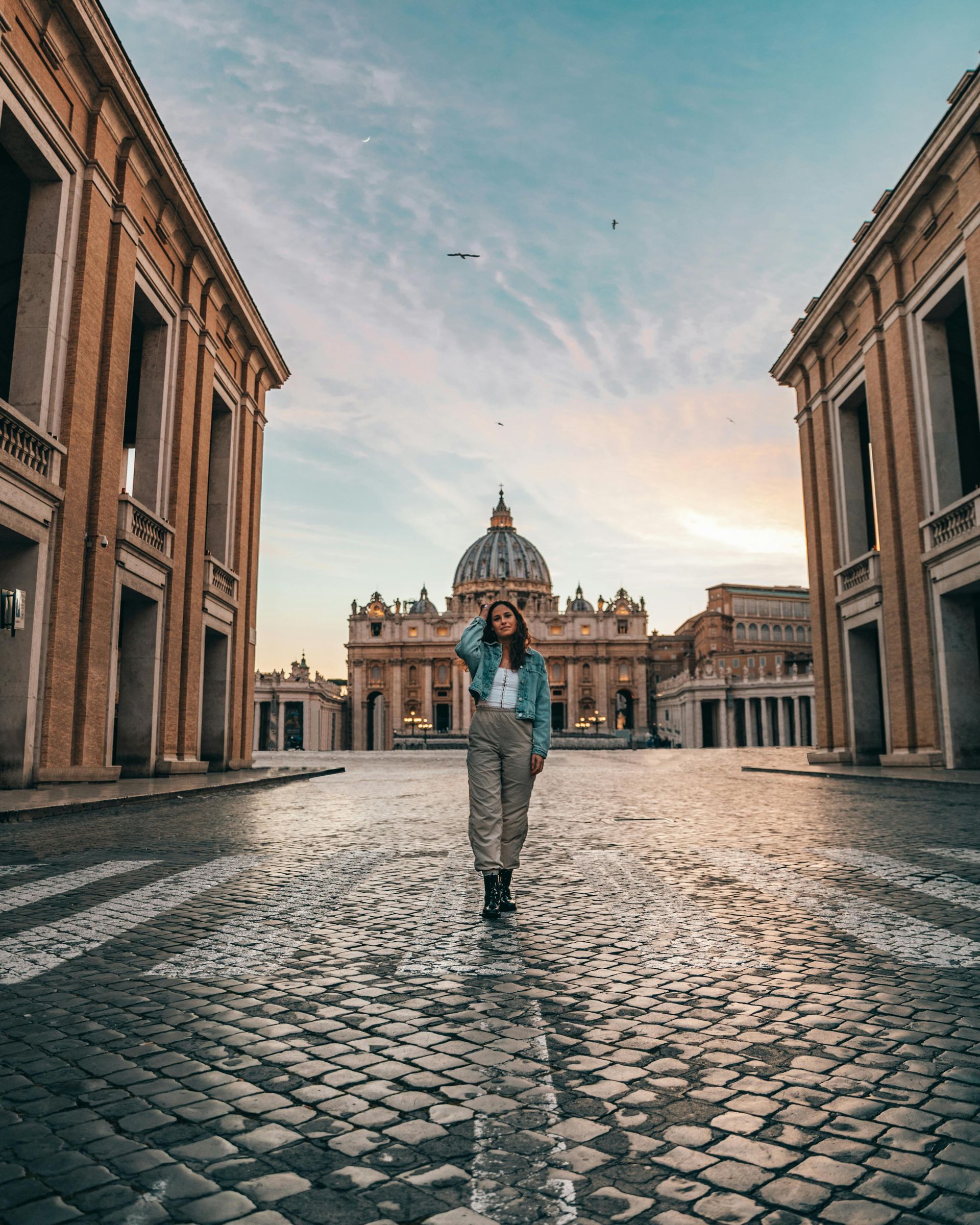 Woman in courtyard, St. Peter's Basilica in background, blue sky, holding hair back, cobblestone pavement.