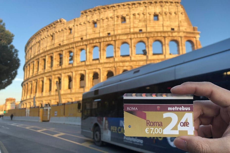 Person holding a Roma 24-hour travel pass in front of the Colosseum and a bus.