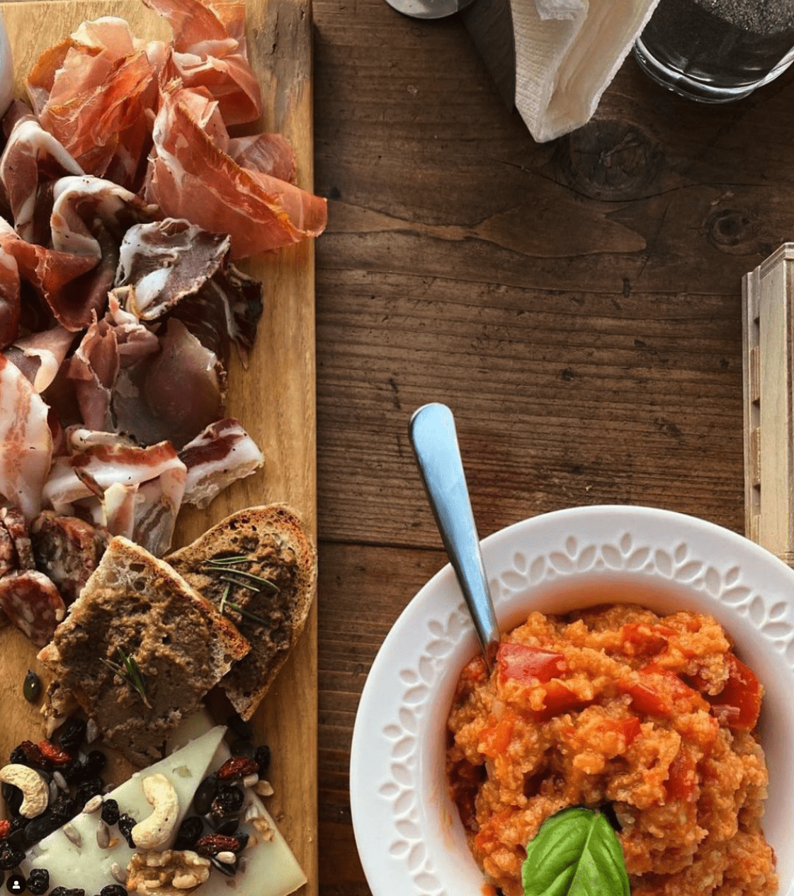 Rustic table with sliced bread, pesto, salami, and a bowl of orange risotto with basil and a blue spoon