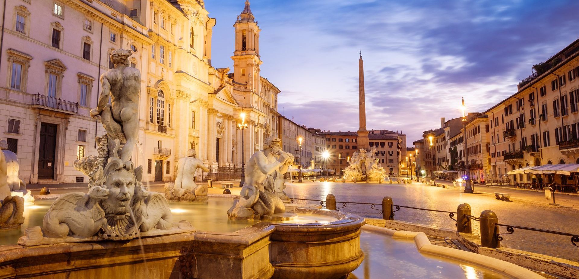 Night view of Piazza Navona in Rome, Italy, with fountains, buildings, and a tall obelisk under a twilight sky.