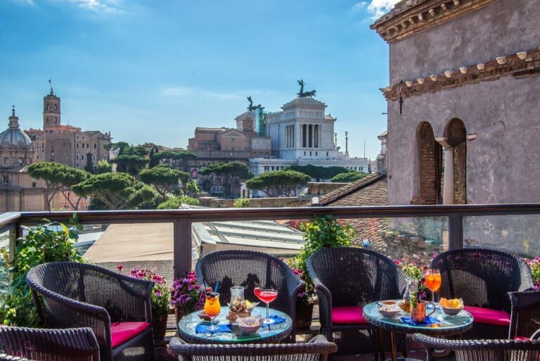 Rooftop terrace with wicker chairs, drinks, and a view of Roman landmarks on a sunny day.