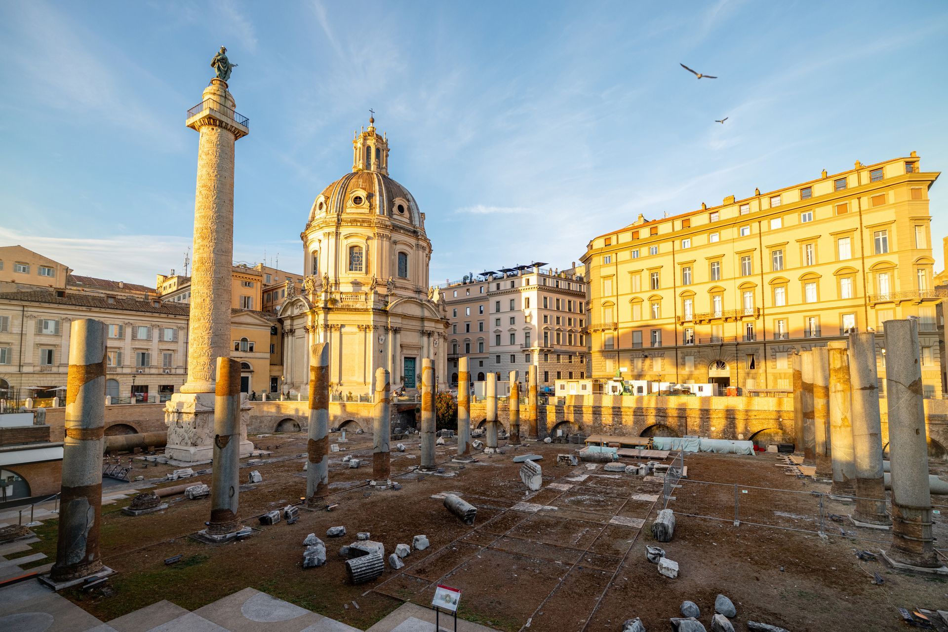 Ruins of ancient Roman forum with Trajan's Column, church, and surrounding buildings under a blue sky.