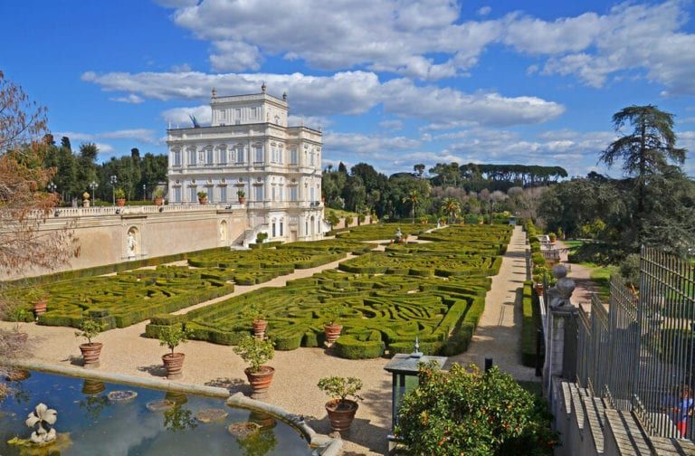 Formal garden with hedges, a large white building, and a blue sky.