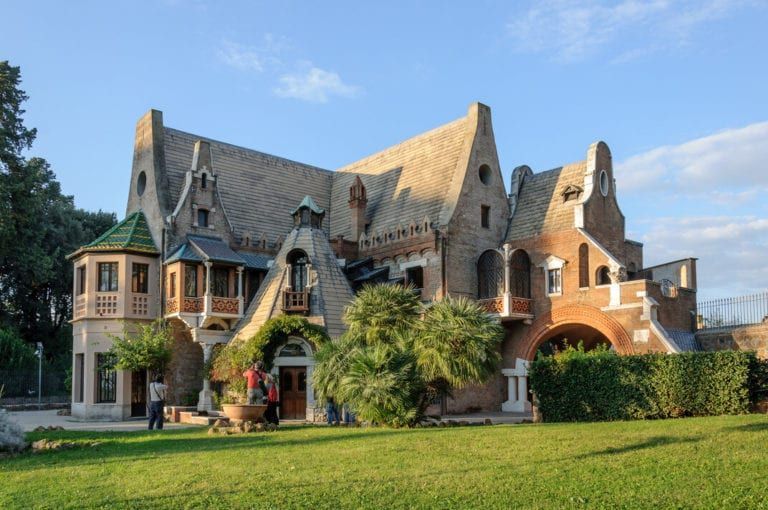 A large, whimsical brick building with gabled roofs and arched windows, set on a green lawn under a blue sky.